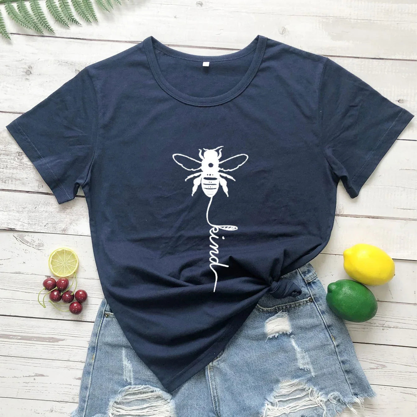 Navy blue t-shirt with white bee and 'be kind' print, paired with ripped denim shorts on wood background, surrounded by fruit for a casual summer look.