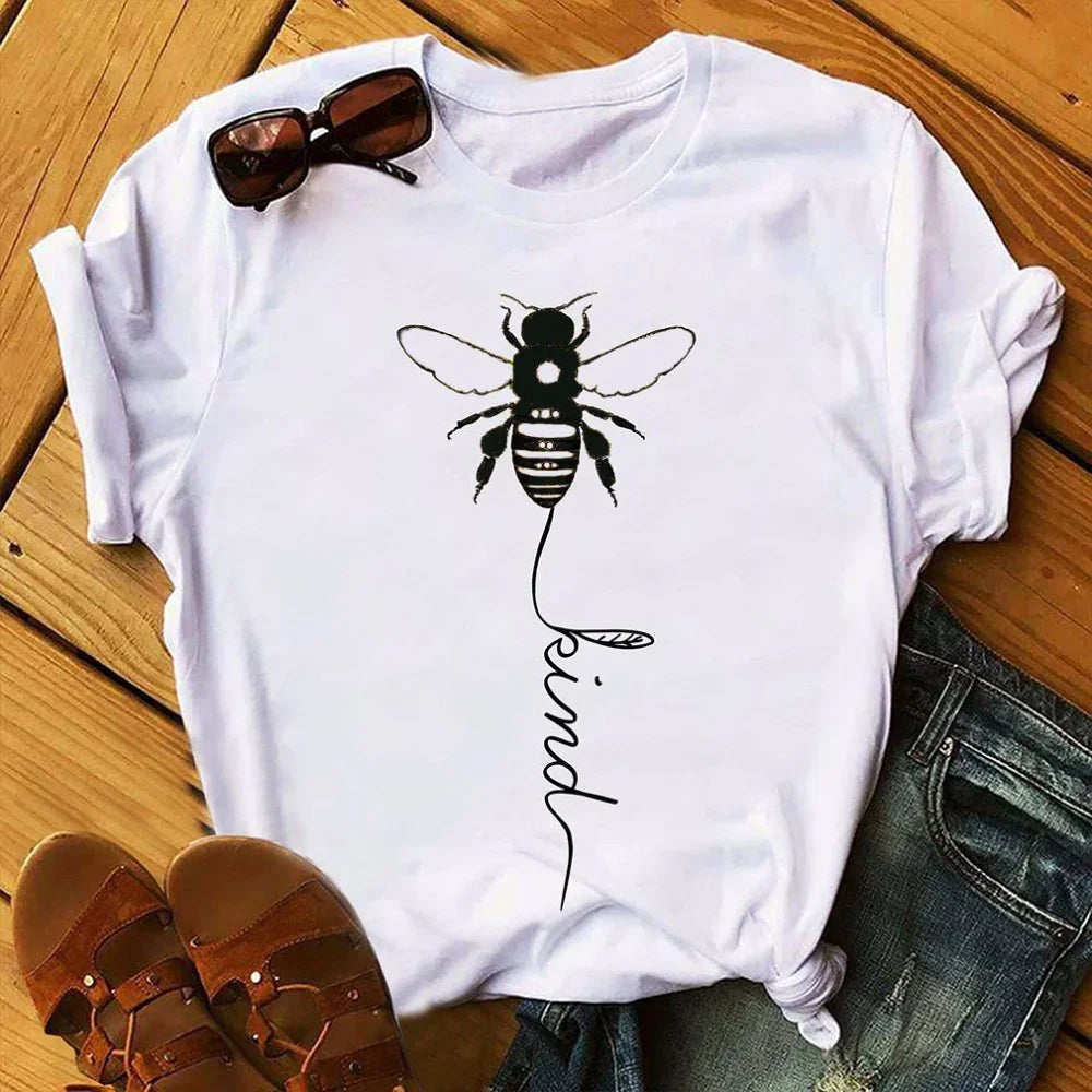 White t-shirt with bee graphic and 'kind' text, styled with sandals and sunglasses on wooden floor.