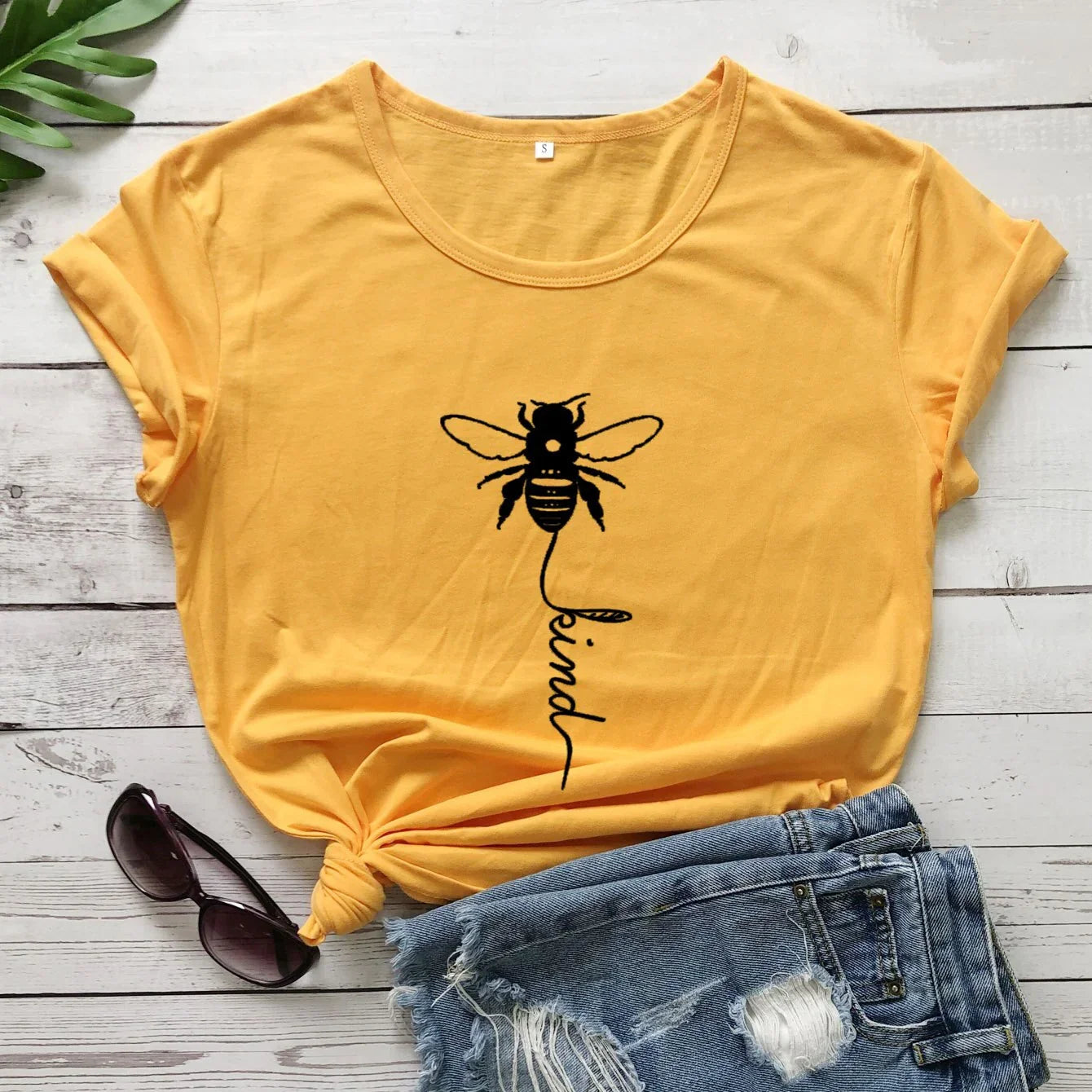 Yellow t-shirt with bee graphic and 'kind' text, paired with denim shorts and sunglasses.